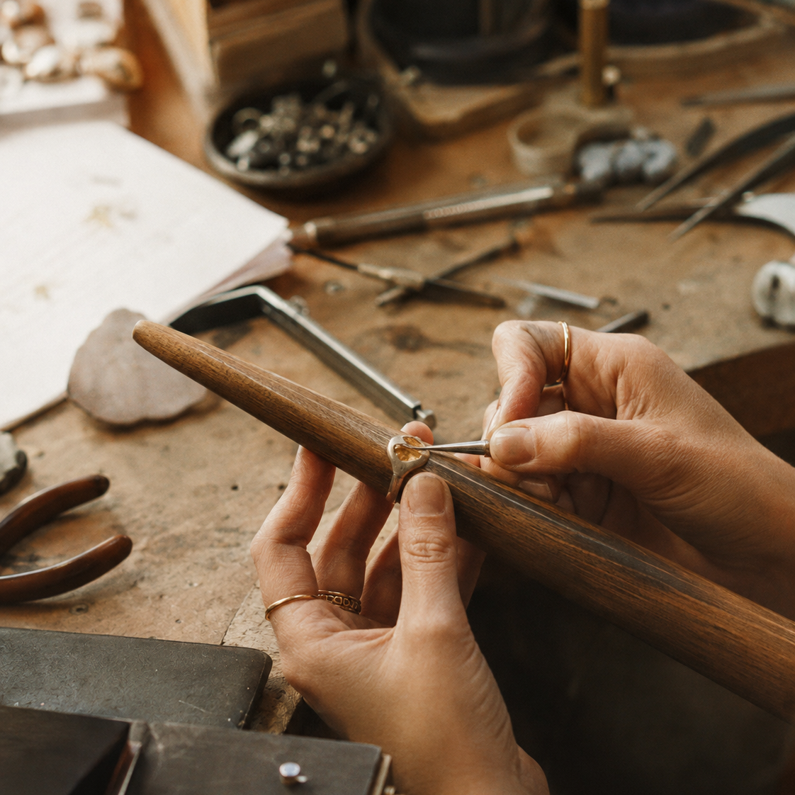 A close-up lifestyle shot of a jeweler's hands using a wooden ring mandrel to shape a gold ring on a rustic, tool-filled workbench.
