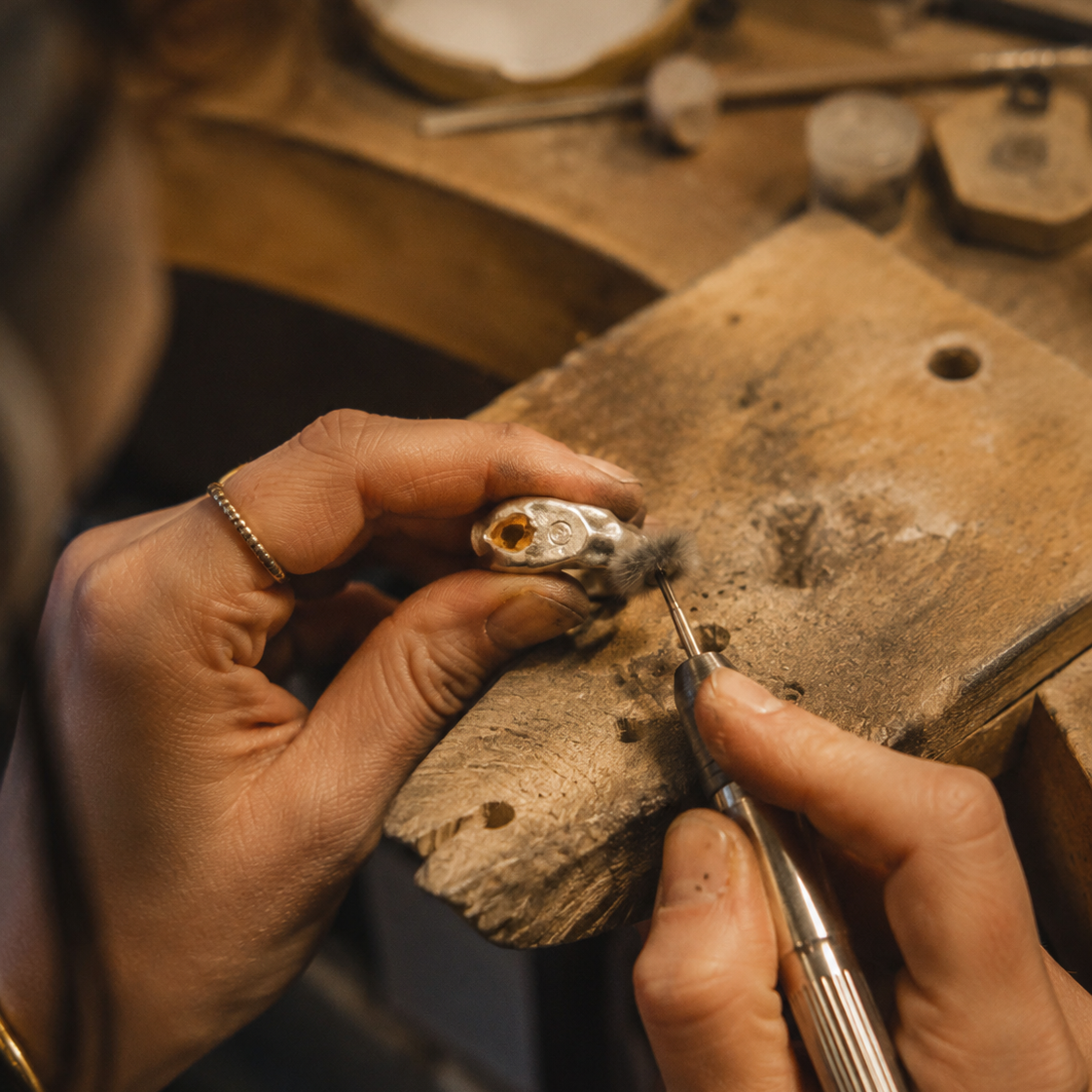 Close-up of a jeweler's hands crafting a small gold ring on a wooden workbench, using precision tools in a warm, focused workshop setting.